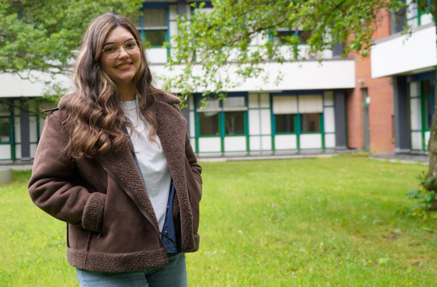 Eine Junge Frau mit langem, welligen Haar steht draußen vor einem Hochschulgebäude. Hinter ihr Wiese, ein Baum und das Gebäude.