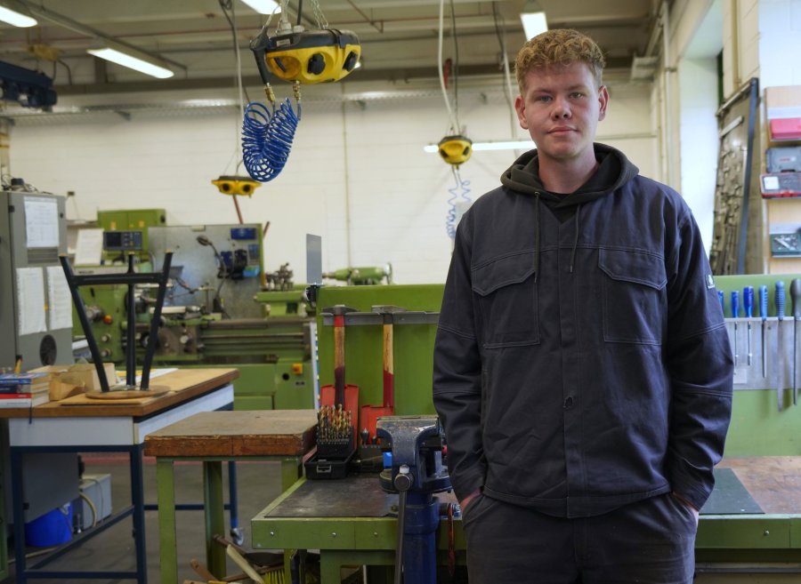 Ein junger Mann steht in einer Werkstatt. Er trägt kurze Haare und Arbeitskleidung, um ihn herum Werkzeige und anderes Equipment.