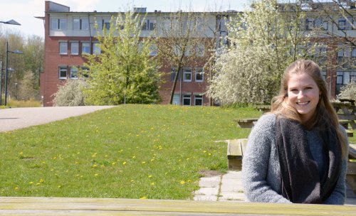 Studentin Nina sitzt an einem sonnigen Tag an einem Tisch auf dem Campus. Im Hintergrund sind eine Wiese und ein Hochschulgebäude zu erkennen.