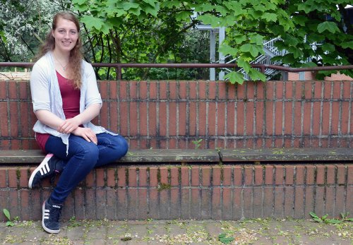 Studentin Jennifer sitzt auf einer Mauer-Bank auf dem Campus, im Hintergrund sieht man grüne Pflanzen.