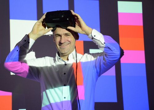 Michael Teistler steht vor einem in Hochschulfarben beleuchteten Hintergrund und setzt sich gerade eine VR-Brille auf.