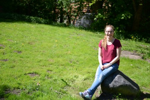 Studentin Sophie sitzt auf einem Findling, der auf einer Wiese steht. Es ist ein sonniger Tag.