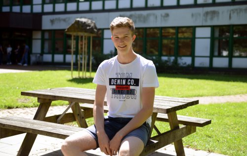 Student Nils sitzt auf einer Holzbank auf einer grünen Wiese. Hinter ihm ist ein Hochschulgebäude zu erkennen. Es ist ein sonniger Tag.