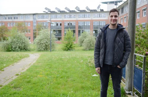 Student Tim steht an einem eher bewölkten Tag auf dem grünen Campus. Rechts von und hinter ihm sind Gebäude zu erkennen. Das Bild wird vom Grün der Wiese auf der er steht, dominiert.