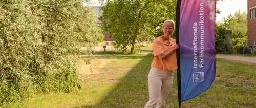 Nele steht auf einer Wiese auf dem Flensburger Campus und präsentiert eine Beachflag mit der Aufschrift "Internationale Fachkommunikation".