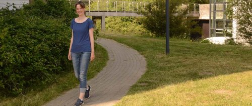 Sarah spaziert über den Flensburger Campus - im Hintergrund ein teilweise verglastes Gebäude und eine Fußgängerbrücke.