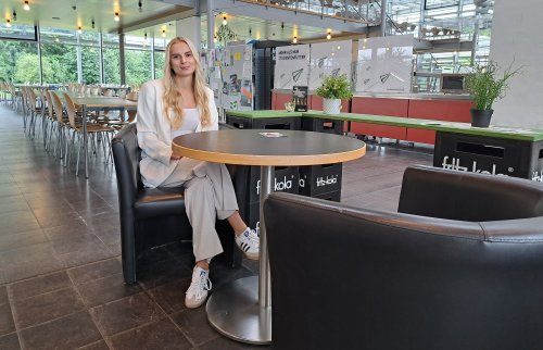 Eine junge Frau im weißen Hosenanzug sitzt in der Flensburger Mensa an einem kleinen Runden Tisch in einem Kunstledersessel. Hinter ihr sieht man Tische und Stühle sowie eine Essenausgabe.