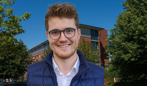 Portraitfoto eines jungen Mannes mit Brille und Dreitagebart, der offen in die Kamera lächelt. Im Hintergrund ein großes Backsteingebäude, Bäume und strahlend blauer Himmel.