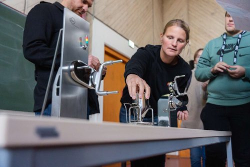 Auf einem Tisch sind mehrere kleine Metallvorrichtungen aufgebaut, eine blonde Frau greift gerade nach einem der Teile. Neben ihr steht ein junger Mann, dem sie gerade etwas erklärt.