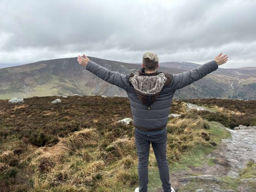 Ein junger Mann steht mit dem Rücken zur Kamera und breitet die Arme nach oben aus. Er steht in einer heide-bedeckten Berglandschaft.