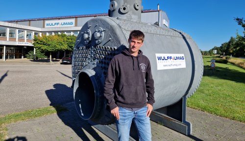 Ein junger Mann im Kapuzenpulli steht auf einem Betriebsgelände vor einem großen Metalltank, auf diesem und dem Gebäude im Hintergrund jeweils der Schriftzug "Wulff & Umag".