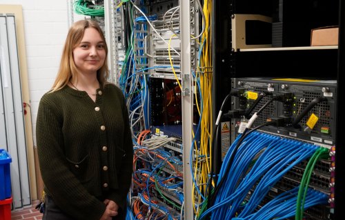 Eien Junge Frau mit langen dunkelblonden Haaren steht in einem Serverraum und blickt in die Kamera. Rechts eine Wand mit vielen Kabeln.