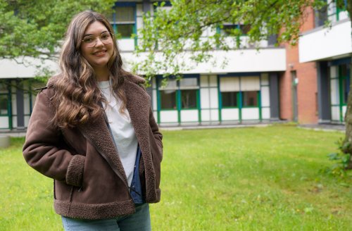 Eine Junge Frau mit langem, welligen Haar steht draußen vor einem Hochschulgebäude. Hinter ihr Wiese, ein Baum und das Gebäude.