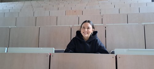 Eine Studentin sitzt in einem Hörsaal und lächelt in die Kamera.