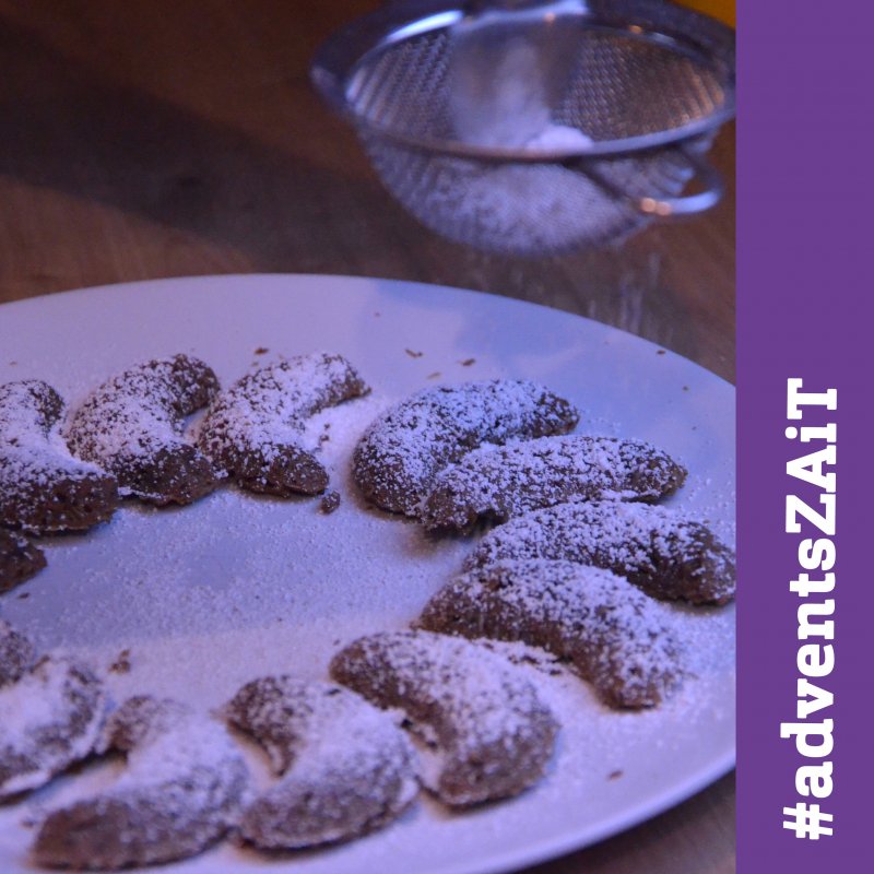 Teller mit Kipferl, oben im Bild ein kleines Sieb aus dem Puderzucker fällt. Rechts ein lila Balken in dem in weiß #adventszait steht.