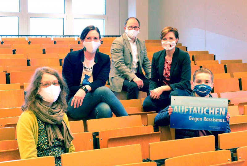 Fünf Menschen sitzen auf Stühlen und Lehnen in einem Hörsaal. Sie tragen Masken, einer hält eien Schild hoch.