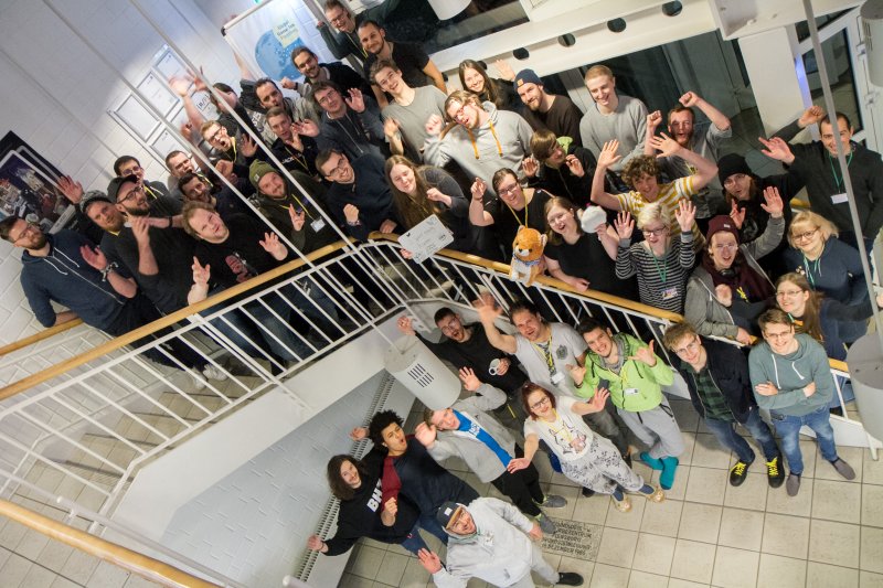 Gruppe Studierender auf und vor einer Treppe aufgereiht. Alle winken in die Kamera.