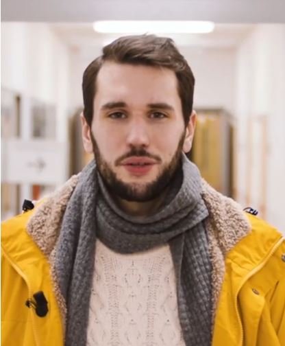 Junger Mann in gelber Jacke in einem Flur. Er sieht direkt in die Kamera.