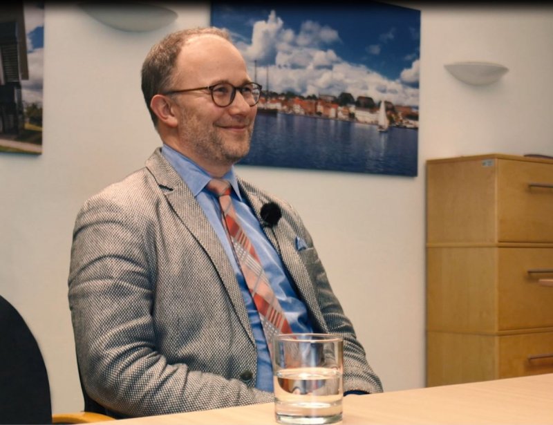 Christoph Jansen sitzt in grauem Anzug, blauen Hemd und gemusterter Krawatte an eine Tisch, vor ihm ein Wasserglas. Er lächelt und ist leicht schräg von der Seite aufgenommen.