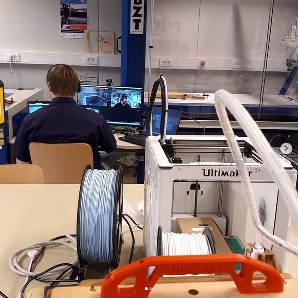 Vorn ein 3D-Drucker aufe einem Arbeitstisch, im Hintergrund sitzt jemand vor einem aufgeklappten Laptop.