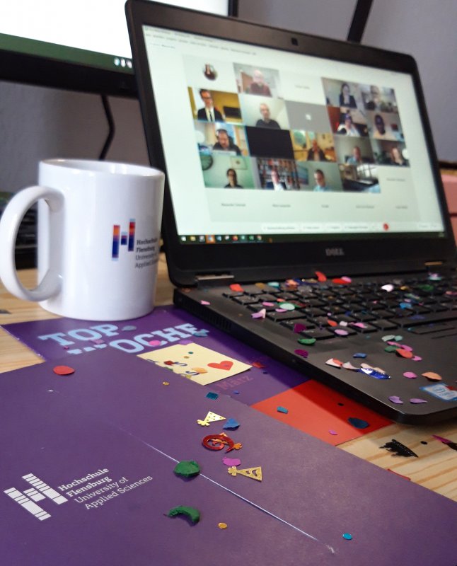 Auf einem Schreibtisch steht ein Laptop, darauf ist eine Videokonferenz zu erkennen. Vorn im Bild Tasse und Unterlagen mit Hochschul-Logo und Konfetti.