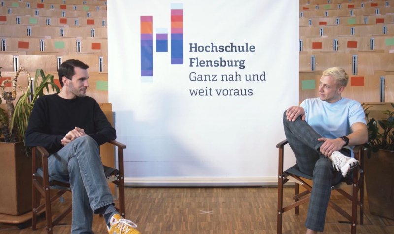Zwei junge Männer in einer Interviewsituation auf Stühlen, dazwischen ein Aufsteller mit Hochschulloge.