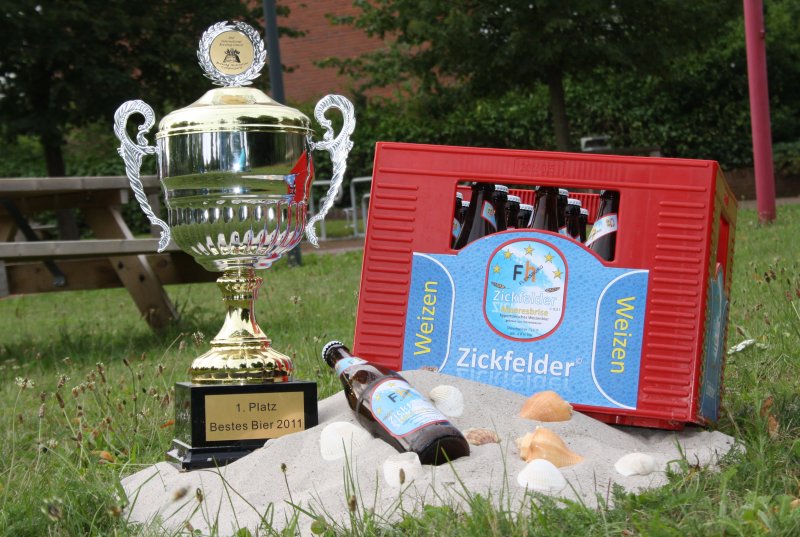 Ein Bierkasten auf einrer Wiese, davor Sand, auf dem eine Bierflasche liegt. Daneben ein Pokal.