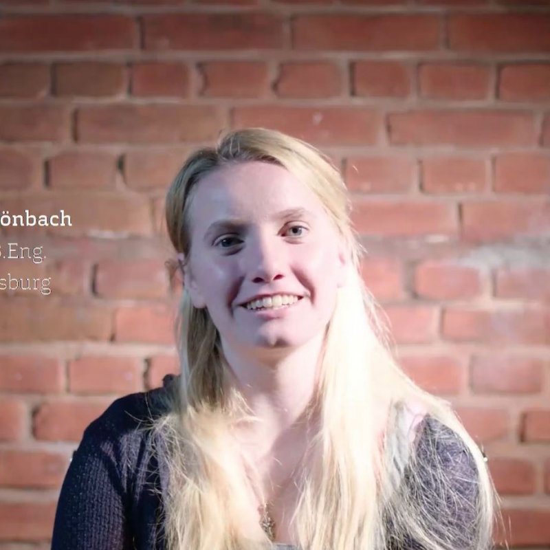 Blonde junge Frau vor einer Backsteinwand. Sie guckt und spricht direkt in die Kamera.