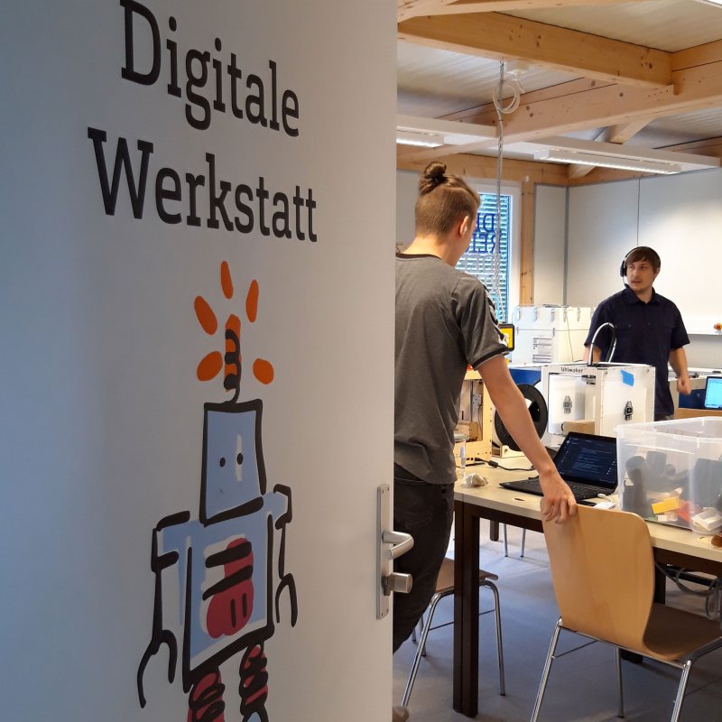Blick durch halbgeöffente Tür auf Werkstatt mit 3D-Druckern und zwei jungen Männern.