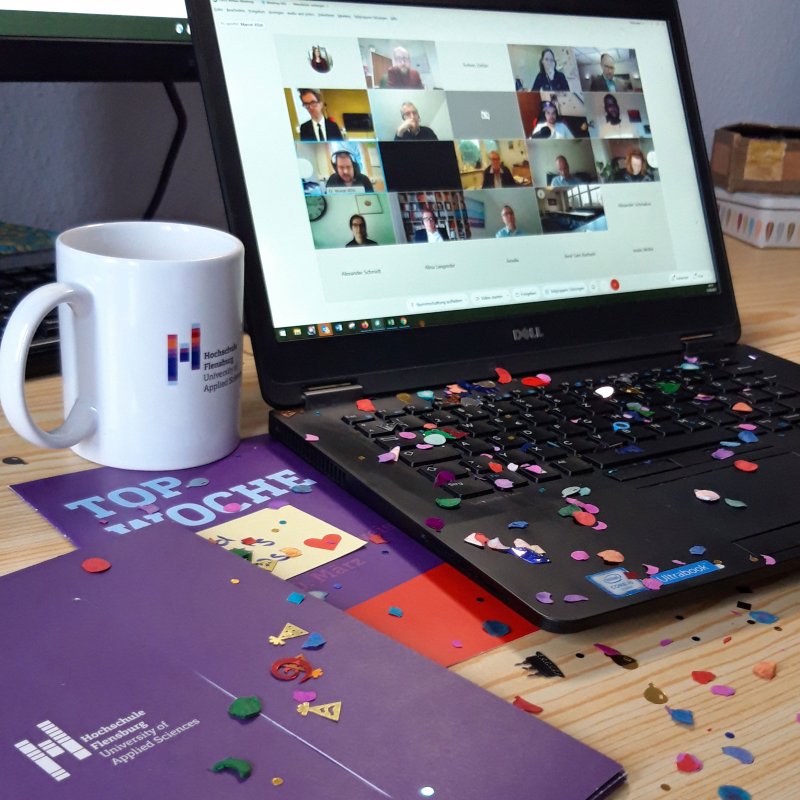 Laptop und Unterlagen in Hochschulfarben auf einem Schreibtisch, darauf ist Konfetti verstreut.