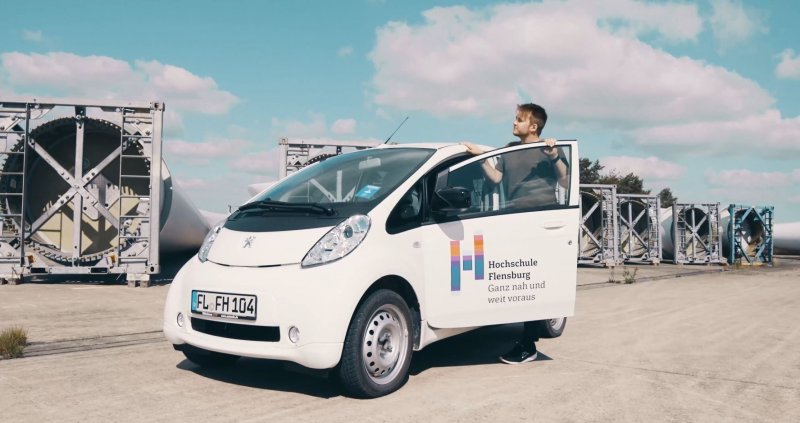 Ein junger Mann steigt aus einem kleinen weißen Auto, hinter ihm leigen Bauteile für Windenergieanlagen.