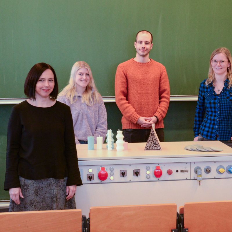 Vier Menschen stehen mit etwas Abstand. Drei stehen hinter einem Tisch, auf dem sie kleine Objekte präsentieren.