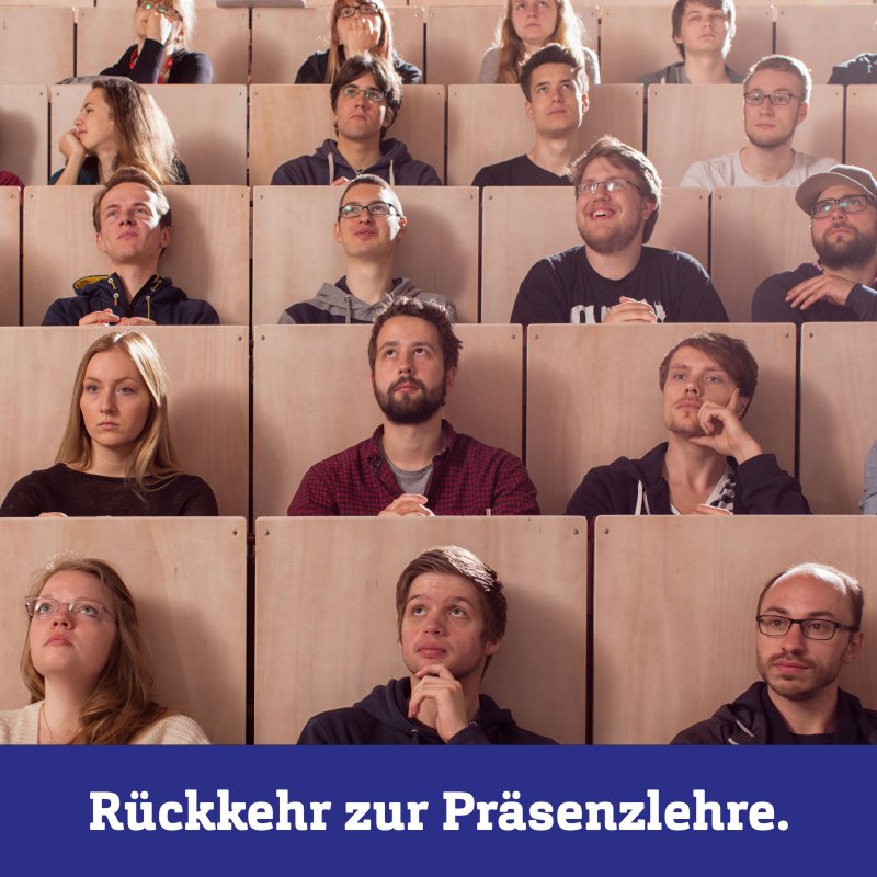 Ein Hörsaal: Mehrere Reihen sind dicht besetzt mit jungen Menschen, die auf etwas schräg über der Kamera blicken. Unten eine blaue Box mit weißer Schrift: Rückkehre in die Präsenzlehre