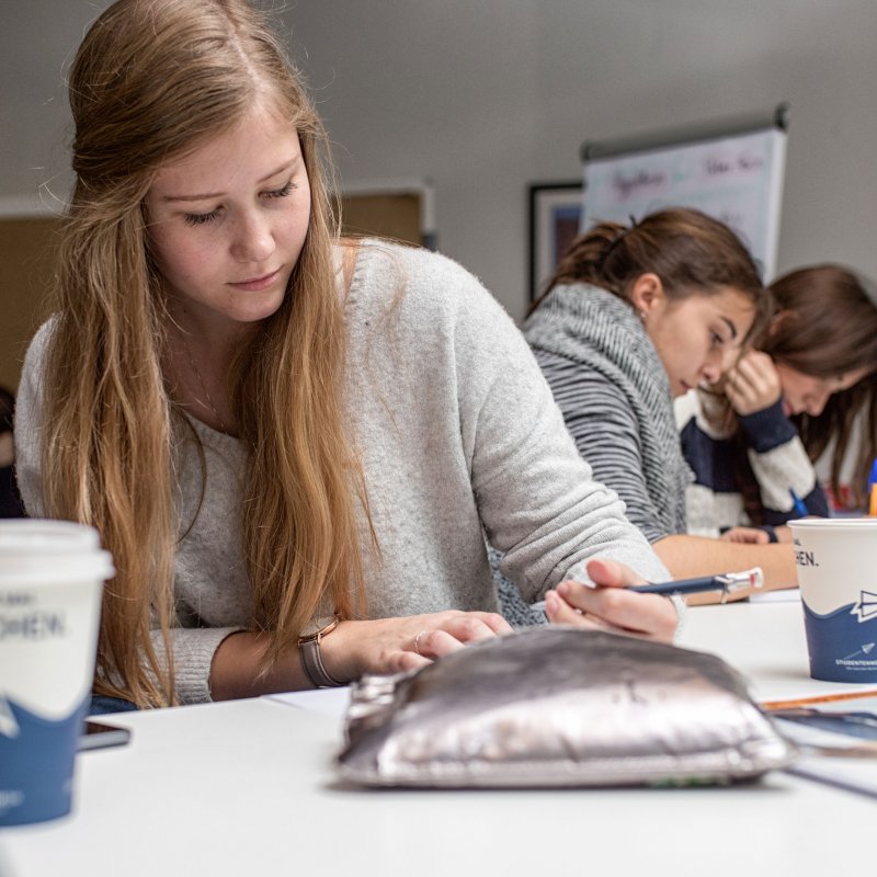 Junge Frau über Unterlagen auf einem Tisch gebeugt. Im Hintergrund sind weitere Menschen zu erkennen.