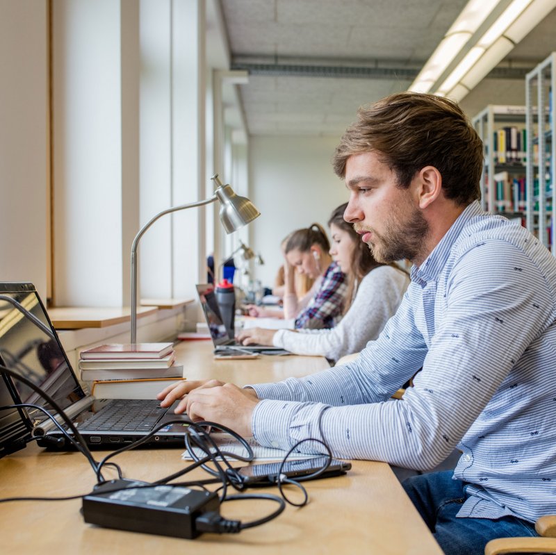 Junger Mann sitzt an einem Arbeitsplatz in der Bibliothek am Laptop, nbenen ihm unscharf andere Menschen an Laptops, im Hintergrund Bücherregale.