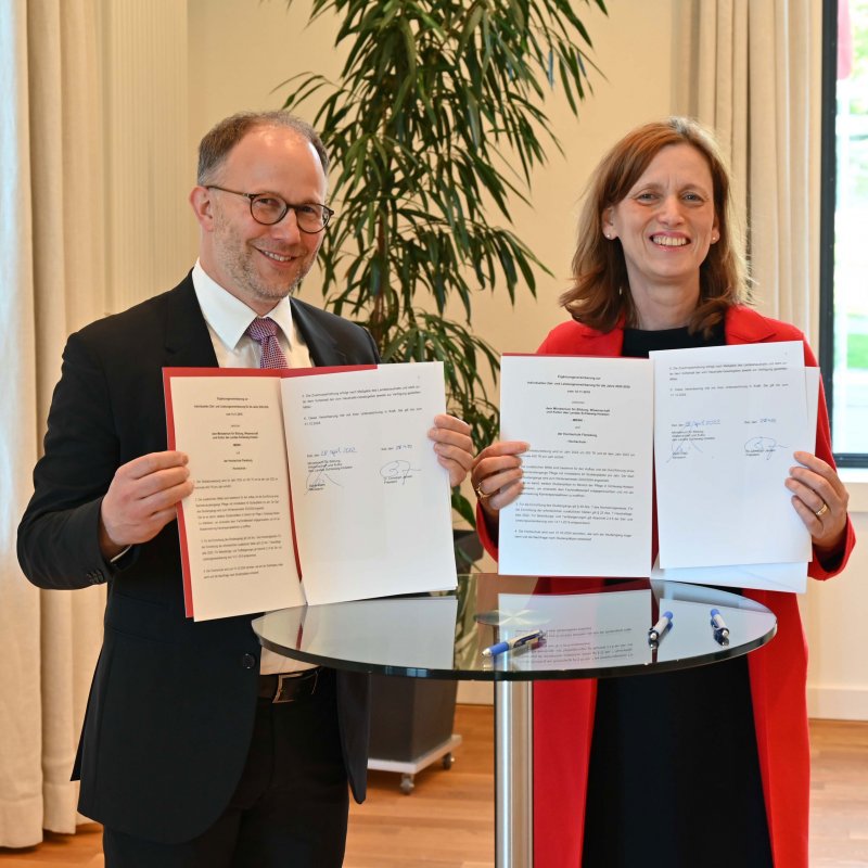 Präsident Christoph Jansen und Ministerin Karin Prien stehen nebeneinander an einem Stehtisch und halten lächelnd Urkunden in die Kamera.
