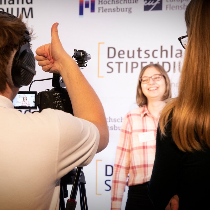 Junger Mann von hinten zu sehen, er hebt den Daumen seiner Hand. Verschwommen sieht man  eine junge Frau, die vor einer Wand mit Deutschland-Stipendium Aufdruck steht.
