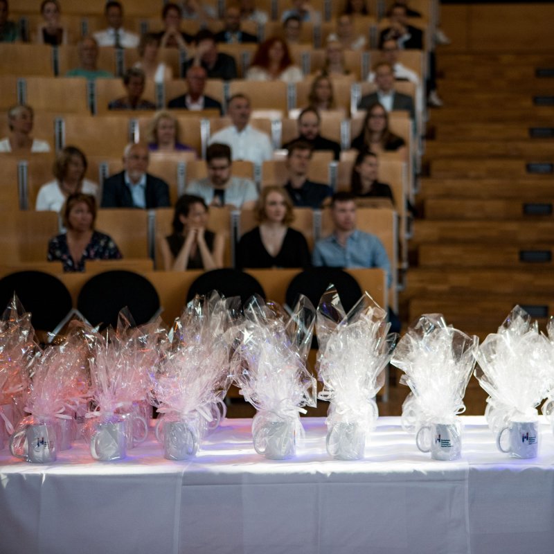 Vorn im Bild ein Tisch mit in Folie verpackten Tassen, dahinter unscharf zu erkennen: Menschen in Sitzreihen in einem Saal.