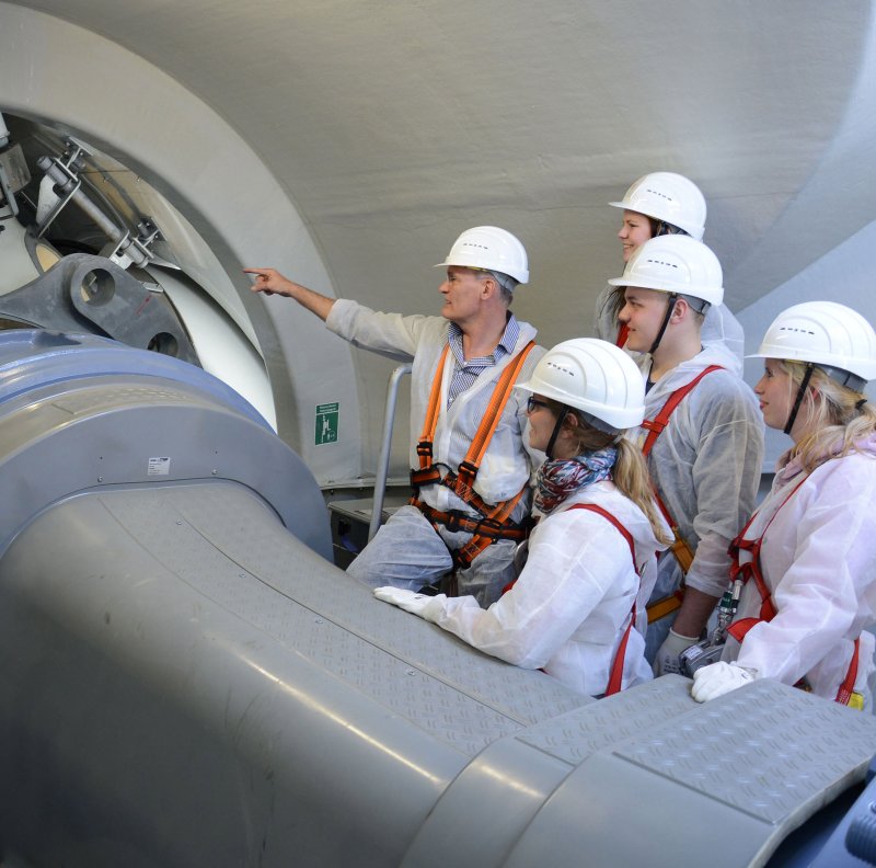 Eine Gruppe Studierender vorm Rotor einer WEA, ein Lehrender zeigt mit der Hand auf einen Teil der technischen Anlage. Alle tragen Schutzhelme.