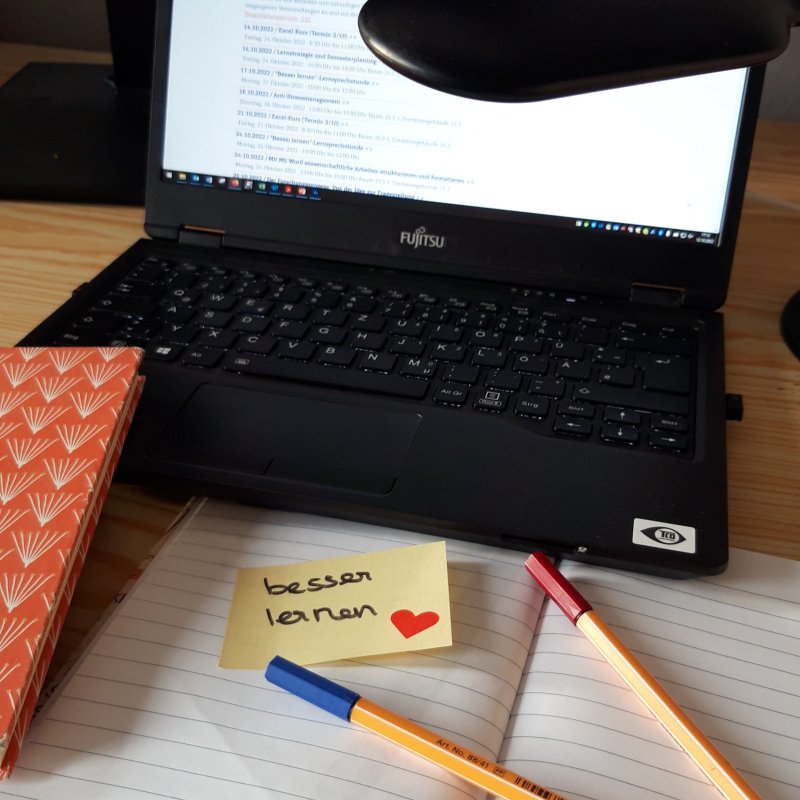 Schreibtisch: Darauf ein Laptop, Stifte und Notizbücher. In der Mitte ein Post It auf dem "besser lernen" steht.