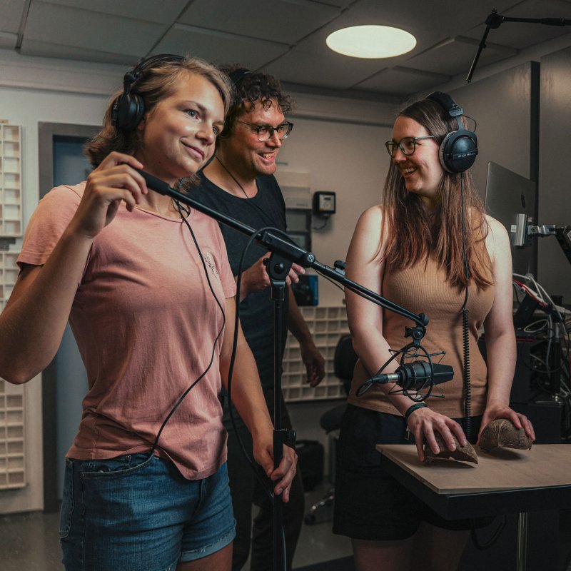 3 Menschen in einem Tonstudio: Links hält eine junge Frau ein Mikrofon, rechts klopft eine junge Frau mit Kokosnusshälften auf eine Oberfläche, dazwischen steht ein Mann. Alle schauen fröhlich.