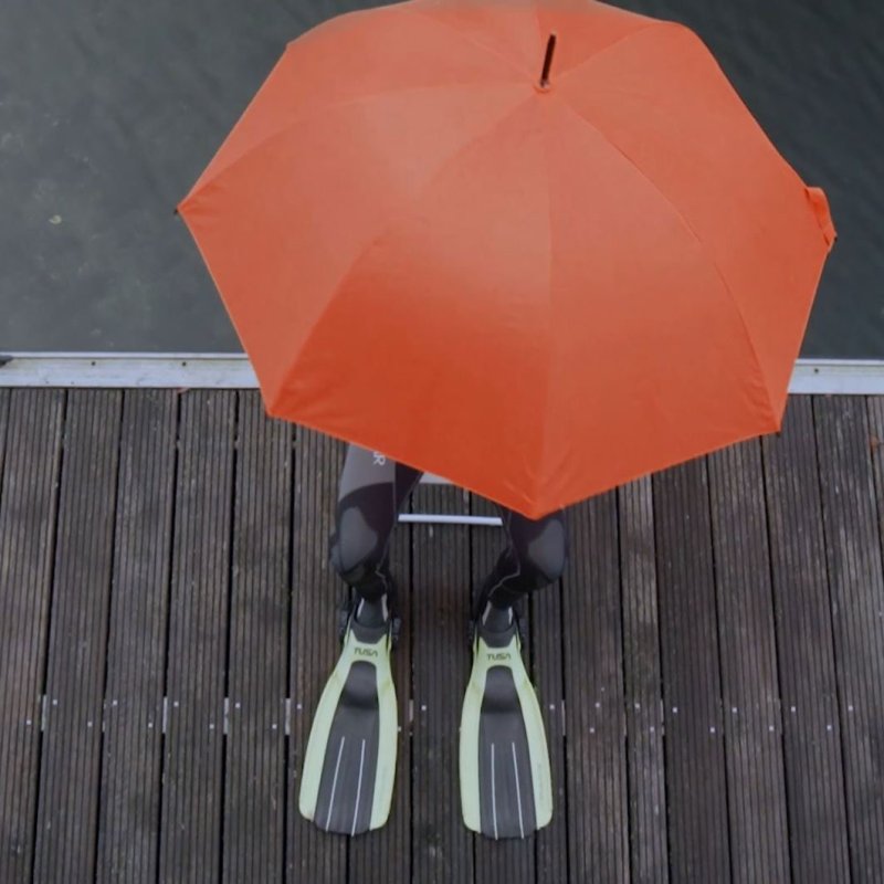 Blick von oben auf einen Menschen im Tauchanzug auf einem Steg sitzend. Er ist zum Großteil von einem aufgespannten orangenen Schirm verdeckt.