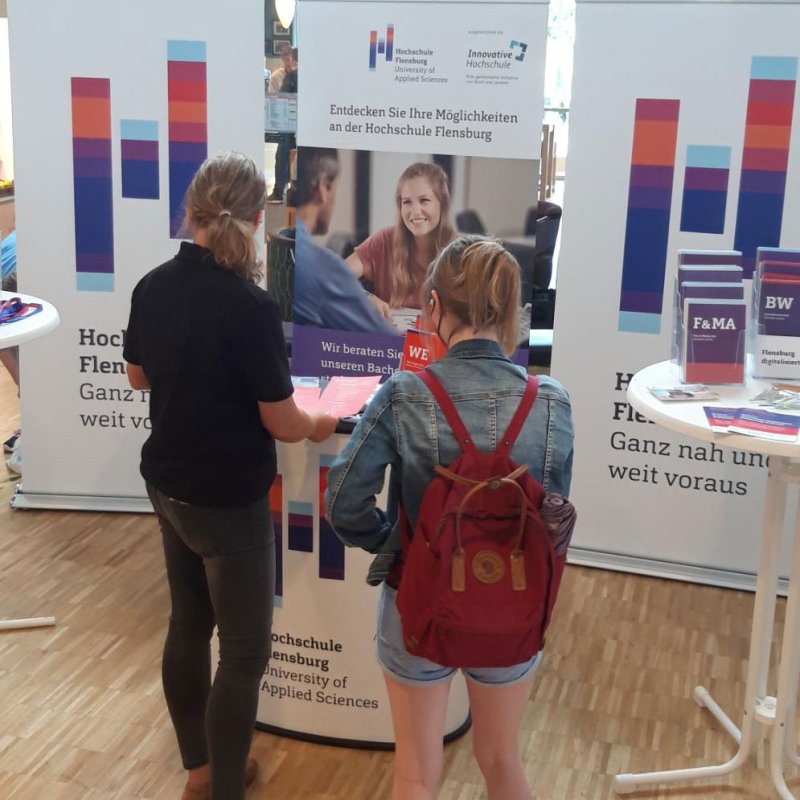 Zwei Menschen an einem Messestand der Hochschule. Sie stehen mit dem Rücken zu Kamera und scheinen Infomaterial anzusehen.