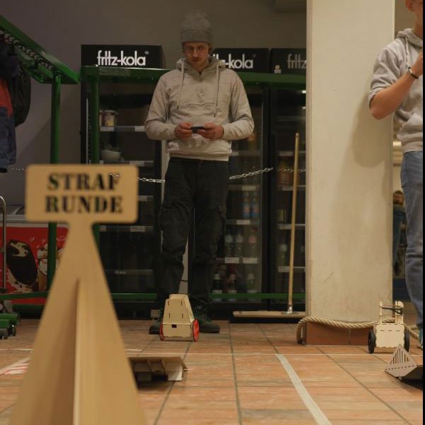 Junger Mann lenkt kleinen Roboter mit Fernbedienung. Vorn im Bild ein Schild, auf dem "Strafrunde" steht.