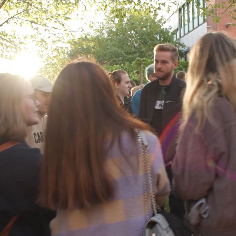 Gruppe junger Menschen stehen einander zugewandt im Kreis und unterhalten sich. Die Sonne scheint und im Hintergrund ist ein grüner Baum zu erkennen.