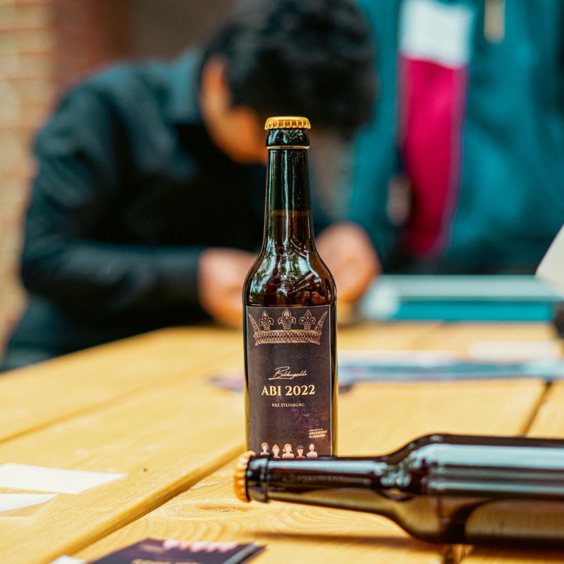 Im Vordergrund steht eine Bierflasche mit selbst designtem Etikett auf einem Tisch, eine andere liegt daneben. Im Hintergrund ist unscharf jemand zu erkennen, der vornüber gebeugt am Tisch arbeitet.