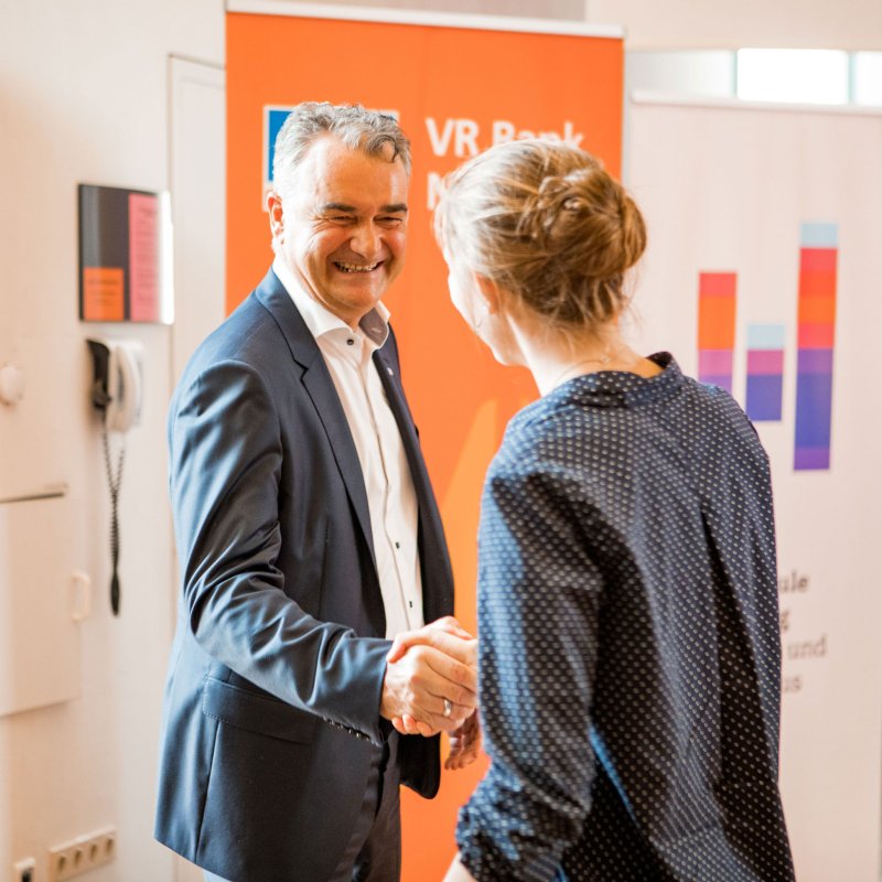 Ein älterer Mann im Anzug schüttelt einer jungen Frau gratulierend die Hand. Sie ist von hinten zu sehen. Im Hintergrund Logos der Hochcshule und der VR Bank.