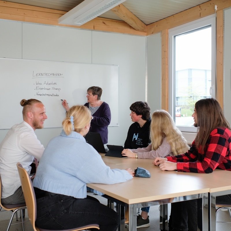 Seminarszene: Studierende sitzen um einen Tisch, vorn steht eine Dozentin am Whiteboard.
