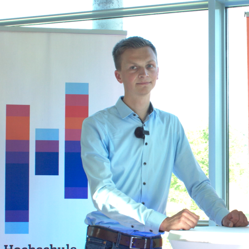 Ein junger Mann im weißen Hemd steht an einem Stehtisch vor einem Roll-Up mit dem Logo der Hochschule Flensburg.