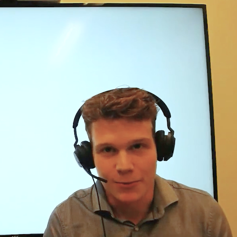 Ein junger Mann im grauen Hemd hat ein Headset auf und spricht direkt in die Kamera. Im Hintergrund ein großer Monitor.